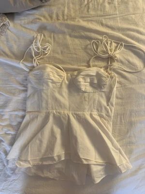 Vit linnetopp med knytband - Säljer en söt vit linnetopp med knytbara axelband och markerade kupor. Toppen har en lätt utsvängd peplumform och är sydd i ett tunt, luftigt material som känns perfekt för varma dagar. Supersnygg att styla med jeans eller kjol!