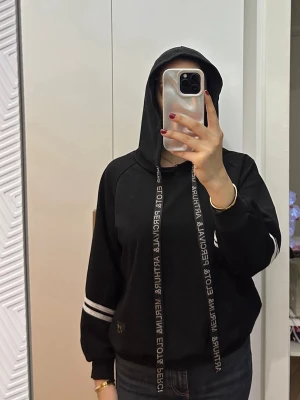 Svart hoodie med vita detaljer - Snygg svart hoodie med huva och långa snören med text. Två vita ränder på ärmarna ger en sportig vibe. Hoodien har ribbade muddar och avslappnad passform, perfekt för kalla dagar. Passar till jeans eller mjukisbyxor.