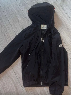 Svart Moncler vindjacka med huva - Snygg svart vindjacka från Moncler med huva och dragkedja framtill. Jackan har Moncler-logga på ärmen och klassiska röd-vit-blå detaljer vid huvan. Tillverkad i lätt och slitstarkt material, perfekt för blåsiga dagar.