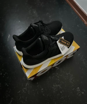 Adidas UltraBoost - Säljer ett par svarta Adidas UltraBoost sneakers med stickad ovandel och vit Boost-sula. Skorna har klassisk snörning och diskret logga på plösen. Perfekta för dig som gillar stilrena och bekväma sneakers med modern design.