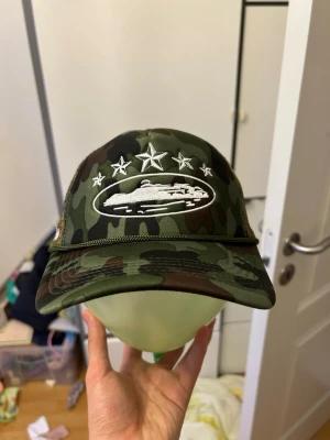 Camo keps med broderade stjärnor - Säljer en grön camouflagemönstrad keps med vit brodyr framtill av ett fordon och fem stjärnor. Sidan har mesh-tyg och fler broderade stjärnor. Justerbar baktill för perfekt passform. Perfekt för dig som gillar streetwear och unika detaljer.