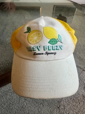 Gul och vit keps med citronmotiv - Säljer en vit och gul truckerkeps med mesh på sidorna och tryck framtill med citroner och texten 'Easy Peezy Lemon Squeezy'. 