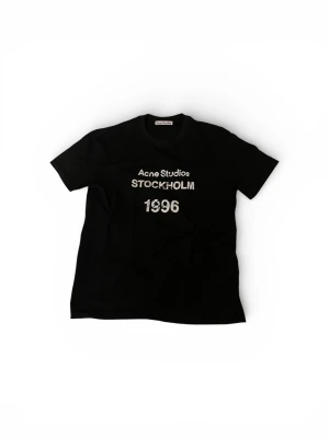 Svart Acne Studios t-shirt 1996 - Snygg svart t-shirt från Acne Studios med tryck i vitt framtill där det står 'Acne Studios STOCKHOLM 1996'. Klassisk rund hals och korta ärmar. Perfekt basplagg med en clean och modern vibe. Hör av dig vid fundering!