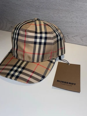 Beige keps med Burberry-rutor - Säljer en klassisk beige keps från Burberry med det ikoniska rutmönstret i svart, vitt och rött. Kepsen har böjd skärm och justerbar rem bak. Tillverkad i ett mjukt och slitstarkt material som ger en clean och trendig look.