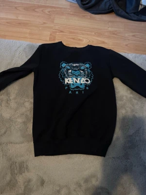 Svart Kenzo sweatshirt med tiger - Säljer en svart sweatshirt från Kenzo med det ikoniska tigerhuvudet broderat i blått och vitt på bröstet. Tröjan har rund halsringning och långa ärmar. Perfekt för dig som gillar streetwear och vill sticka ut med en snygg logga.