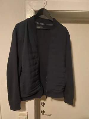 Mörkblå bomberjacka från Fynch Hatton - Snygg mörkblå bomberjacka från Peak Performance med quiltad framsida och ribbade muddar. Jackan har en klassisk bomberkrage och dragkedja framtill. Perfekt för dig som vill ha en stilren och enkel look.