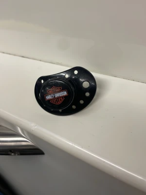 Svart napp Harley-Davidson - Cool svart napp med Harley-Davidson-logga på framsidan. Nappen är rund med flera små hål runtom och är tillverkad i plast. Perfekt accessoar för den lilla motorcykelfantasten eller som rolig present. One in a kind napp