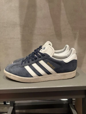 Adidas Gazelle blå/vita sneakers - Klassiska Adidas Gazelle sneakers i blå mocka med vita detaljer och de ikoniska tre ränderna på sidorna. Skon har vit sula, vita hälpartier och blå snörning. Guldtexten 'GAZELLE' syns på sidan. Perfekt för en stilren och sportig look.