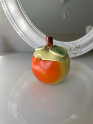 Keramikburk formad som äpple - Charmig liten burk med lock, utformad som en frukt (äpple/persika) i fina mjuka färger – orange, gul och grön med dekorativt blad och skaft. Perfekt som sockerskål, godisburk eller bara som fin retrodekoration.  Troligen tillverkad som exportporslin (“imported”) omkring mitten av 1900-talet. . Burken är märkt med ”Import” undertill. Det finns nagg på kanten på bilden 6