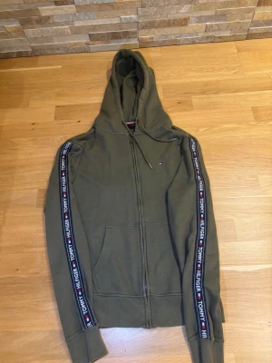 Olivgrön hoodie från Tommy Hilfiger - Olivgrön hoodie med dragkedja från Tommy Hilfiger. Snygga logoband längs ärmarna med texten 'Tommy Hilfiger' och flagga. Huva med dragsko och känguruficka framtill. Perfekt för en avslappnad streetstyle-look.