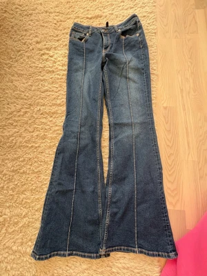 Blå bootcut jeans - Säljer ett par blå bootcut jeans med markerade kontrastsömmar längs benen. Jeansen har klassisk femficksdesign och är tillverkade i denim. Perfekt för dig som gillar retrostil och vill ha ett par jeans med snygg passform och utsvängda ben.