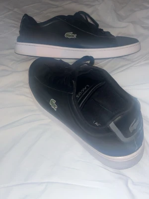 Svarta Lacoste sneakers med krokodillogo - Snygga svarta sneakers från Lacoste med klassisk grön krokodillogo på sidan. Skorna har svarta skosnören, vit sula och är tillverkade i läder. Perfekt för dig som gillar stilrena och sportiga skor.