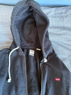 Svart zip-hoodie från Levi's - Svart hoodie med dragkedja från Levi's. Klassisk modell med vit dragsko i huvan, två fickor framtill och liten röd Levi's-logga på bröstet. Perfekt för en avslappnad streetstil. Mjuk bomullskänsla och ribbade muddar vid ärmslut och nederkant.
