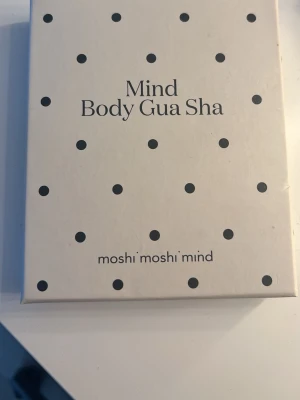 Mind Body Gua Sha från Moshi Moshi Mind - Säljer en Mind Body Gua Sha från Moshi Moshi Mind. Kommer i en vit förpackning med svarta prickar. Perfekt verktyg för massage och ökad blodcirkulation, kan användas på ansikte och kropp. Enkel att rengöra med varmt vatten.