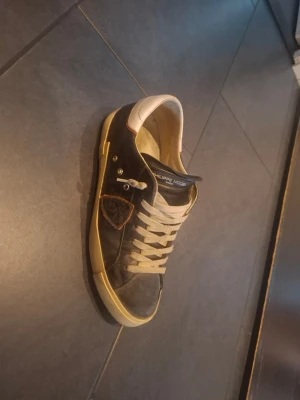 Philippe Model svarta sneakers - Svarta sneakers från Philippe Model med vita snören och vit sula. Skon har en vit hälkappa och en dekorativ sköld på sidan. Tillverkad i läder med klassisk låg profil och diskreta detaljer. Perfekt för dig som gillar stilrena och tidlösa sneakers.