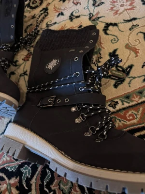 Svarta vinterkängor med snörning - Snygga svarta kängor med grov sula, stickad kant upptill och rejäla snören i svartvitt mönster. Kängorna har metallhakar, spänne på sidan och ett snöflingemärke. Perfekta för kalla dagar och outdoor-äventyr.