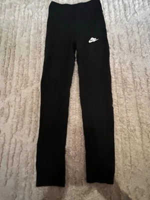 Svarta leggings från Nike - Säljer ett par svarta leggings från Nike med vit logga på vänster höft. De har hög midja och är i stretchigt material som sitter tight. Perfekta till både träning och chill. Klassisk och enkel design som funkar till det mesta.