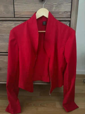 Röd kavaj med dragkedja  - Snygg röd kavaj med modern, figurnära passform och dragkedja framtill. Kavajen har långa ärmar och en öppen krage utan slag. Perfekt för att lyfta vilken outfit som helst med en färgklick.