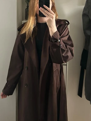 Mörkbrun trenchcoat med knytskärp - Stilren mörkbrun trenchcoat i mjukt material med klassisk dubbelknäppning, bred krage och knytskärp i midjan. Jackan har axelklaffar, fickor och justerbara band vid ärmsluten för extra detaljerad look. Endast använd en gång, från Gina tricot i storlek S💕