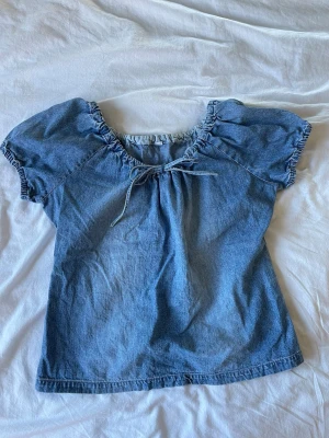 Blå denimtop med knyt och puffärm - Söt blå denimtop med korta puffärmar och knyt i halsringningen. Toppen har en avslappnad passform och elastiska detaljer vid ärmslut och hals. Perfekt för en trendig och avslappnad look.