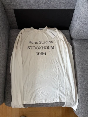 Acne studios long sleeve - Säljer en vit långärmad t-shirt från Acne Studios med tryck 'Acne Studios STOCKHOLM 1996' på bröstet. Klassisk rund halsringning och mjukt material som känns skönt mot huden. Perfekt för dig som gillar minimalistisk och stilren design.