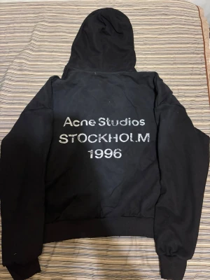 Acne studios hoddie - Säljer en svart hoodie från Acne Studios med texten 'Acne Studios STOCKHOLM 1996' i vitt på ryggen. Klassisk passform med huva och ribbade muddar vid ärmslut och nederkant. Perfekt för dig som gillar stilrena och trendiga plagg.