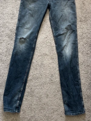 REPLAY Anbass jeans med litet hål på knät  - Säljer ett par blå replay med slitningar och ett hål på vänster knä. Säljer jeansen för jag tycker dom är för små för mig. Skick 7/10. Den lilla replay biten som sitter vid fickan har gått lite sönder. Annars inga större fel 