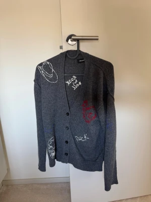 Grå kofta från Zadig & Voltaire - Grå kofta med knappar från Zadig & Voltaire. Koftan har coola broderade detaljer i vitt, rött och blått, bland annat text och motiv på bröstet och ärmen. Mjuk och skön med avslappnad passform och ribbade muddar.