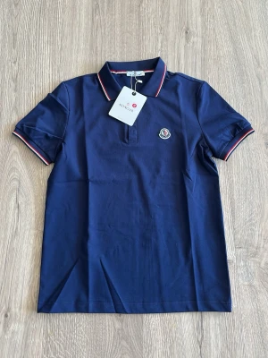 Marinblå pikétröja från Moncler - Snygg marinblå pikétröja från Moncler med klassisk krage och korta ärmar. Tröjan har Moncler-logga på bröstet och röda samt vita detaljer på krage och ärmslut. Perfekt för en stilren look.