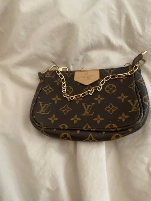 Mini brun handväska  - Brun mini handväska med monogram mönster, guldkedja och röd tyg inuti. Säljer endast väskan, inte assceoarer runt den. Den passar till allt för färgen är så neutral. Exempel på vad som får plats i väskan: telefon, hemnycklar, bilnycklar,läppglans, plånbok. Pepparsprej, ifall en man börjar irritera dig ute med tjejerna. Några små trådar sticker ut men inget som inte går att fixa själv om man vill sitta och pyssla med väskan en liten stund.  Skriv till mig om du har frågor!  ❤️