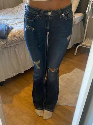 Mörkblå bootcut jeans med slitningar - Snygga mörkblå bootcut jeans med slitningar och dekorativa vita sömmar på bakfickorna. Jeansen har låg midja och klassisk femficksdesign. Perfekta för en trendig och avslappnad look.