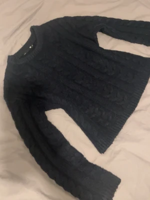 Mörkblå kabelstickad tröja  - Säljer en mörkblå kabelstickad tröja från lager med rund halsringning och långa ärmar. Tröjan har ett klassiskt flätmönster och är gjord i mjukt material som passar perfekt när det är kyligt ute. Ribbstickade muddar vid ärmslut och nederkant.