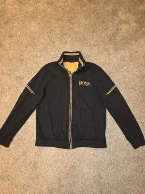 Svart ziptröja från Hugo Boss - Cool svart ziptröja från Hugo Boss med gulddetaljer och logga på bröstet. Tröjan har hög krage med BOSS-tryck, dragkedja och två fickor. Perfekt för dig som gillar stilrena och exklusiva plagg.