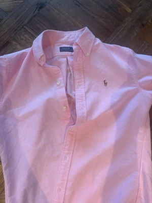 Rosa skjorta från Polo Ralph Lauren - Klassisk ljusrosa skjorta från Polo Ralph Lauren med kort ärm och button-down krage. Skjortan har broderad logga på bröstet och knappar framtill. Perfekt för en stilren och avslappnad look.