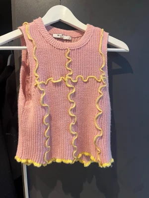 Rosa stickad väst från NA-KD - Supersöt rosa stickad väst från NA-KD med vågiga gula detaljer längs kanterna och framtill. Västen har en ribbad struktur och är ärmlös med rund hals. Perfekt för att addera färg till din outfit!