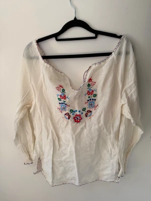 Vit boho-blus med broderi - Superfin vit blus i boho-stil med färgglada broderier framtill. Blusen har v-ringning och långa ärmar med lätt rynkade detaljer. Materialet känns luftigt och mjukt, perfekt för en avslappnad look. Broderierna går i rött, blått, grönt och turkost.