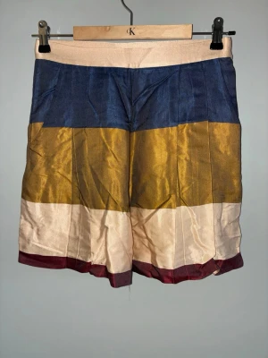 Randiga shorts från H&M i satin - Säljer ett par färgglada randiga shorts från H&M i satinliknande material. Shortsen har breda blockränder i marinblått, gult, beige och vinrött. De har hög midja och en lös passform som ger en chill vibe.