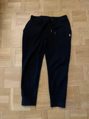 Svarta mjukisbyxor från Polo Ralph Lauren - Svarta mjukisbyxor från Polo Ralph Lauren med klassisk vit logga broderad på ena benet. Byxorna har dragsko i midjan och muddar vid bensluten. Perfekta för en avslappnad och stilren look.