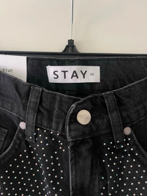 Svarta jeans med strass från STAY - Säljer ett par svarta jeans från STAY i storlek XS. Jeansen har raka ben och är täckta med små glittrande strassdetaljer över hela framsidan. Klassisk femficksmodell med hög midja och knappgylf. Perfekta för dig som vill sticka ut!