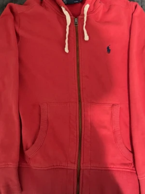 Röd hoodie från Ralph Lauren - Snygg röd hoodie från Ralph Lauren med dragkedja, kängurufickor och vit dragsko i huvan. Klassisk blå logga broderad på bröstet. Mjuk bomullskvalitet och ribbade muddar vid ärmslut och nederkant. Perfekt för en avslappnad och stilren look.