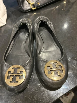Svarta ballerinas från Tory Burch - Snygga svarta ballerinaskor i läder från Tory Burch med elastisk häl och ikonisk guldfärgad logga framtill. Skorna har rund tå och klassisk design som passar till många olika outfits.
