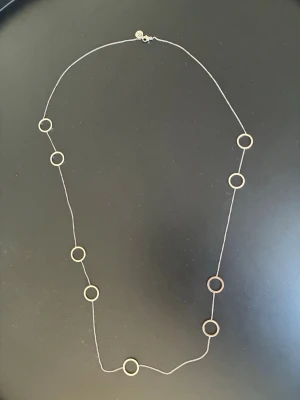 Edblad - långt halsband med ringar - Stilrent långt halsband i silverfärgad metall med flera runda ringar längs kedjan. Kedjan är tunn och smidig, och ringarna är jämnt placerade för en snygg look. Från Edblad. Oanvänt.