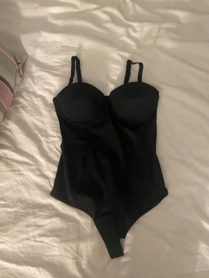 Svart body med bygel och justerbara band - Snygg svart body med vadderade kupor och bygel för extra stöd. Justerbara axelband och klassisk siluett. Materialet är slätt och stretchigt för en bekväm passform. Perfekt att styla under kläder eller som statement-plagg.