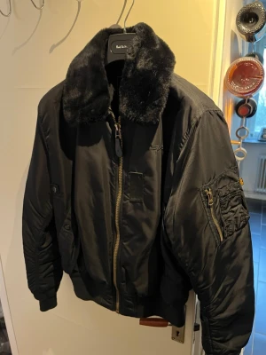 Svart bomberjacka Alpha Industries B-15 - Säljer en svart bomberjacka från Alpha Industries med en stor, mjuk päls-krage. Jackan har dragkedja i guld, fickor på ärmen och ribbade muddar. Klassisk militärinspirerad look som är både stilren och cool. Modellen är B-15