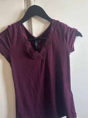 Vinröd topp med spetsdetalj H&M - Vinröd topp från H&M med v-ringning och spetsdetaljer längs halsringningen. Kortärmad och i mjukt bomullsmaterial. Perfekt för dig som gillar en enkel men ändå söt stil.