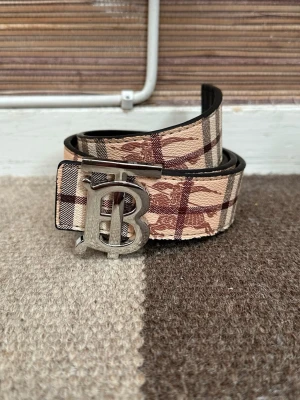 Burberry rutigt bälte med B-spänne - Snyggt bälte från Burberry med klassiskt rutigt mönster i beige, brunt och svart. Bältet har ett stort silvrigt B-spänne och är tillverkat i skinn med tygdetaljer. Perfekt accessoar för att lyfta din outfit.