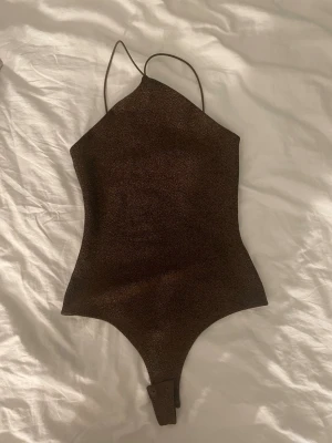Brun glittrig body med smala band - Snygg brun body med glittrigt tyg och tunna axelband. Bodyn har hög skärning vid benen och knäppning i grenen. Materialet ser stretchigt ut och ger en trendig look. Perfekt för dig som vill ha något extra till outfiten.