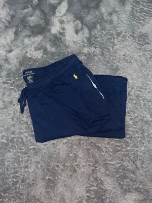 Polo Ralph Lauren Mjukisbyxor/Loungewear - supermjuka loungewear-byxor från Polo Ralph Lauren i marinblått. De har dragsko i midjan för bästa passform och den ikoniska hästen broderad i gult på höften. Byxorna har raka ben och är gjorda i en mjuk, skön bomullsblandning (ser ut som tunnare sweat- eller jersey-material). Perfekta för att slappa hemma i eller som pyjamasbyxor! 
