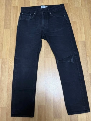 Svarta jeans med slitning från 157 - Säljer ett par svarta jeans från 157 med rak passform och en snygg slitning över ena knät. Klassisk femficksmodell och normal midja. Jeansen är gjorda i ett mjukt denimtyg som ger en avslappnad look. Storlek W32 L30