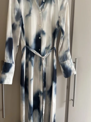 Långklänning i blåvitt mönster h&m - Snygg långklänning med knappar framtill och bälte i midjan. Klänningen har ett unikt blåvitt mönster, lång ärm med bred manschett och klassisk krage. Materialet känns glansigt och mjukt, troligen polyester eller satin.